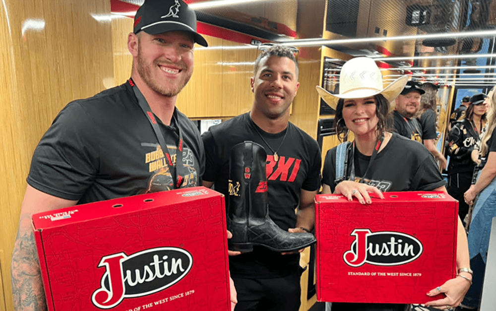 Alltroo Co-Founder Kyle Rudolph on the left holding a red Justin boot box, posing with 23XI Racing Driver Bubba Wallace, who is holding a black custom boot wearing a black t-shirt, standing next to Jenna Paulette on the right who is also holding a red Justin boot box. 